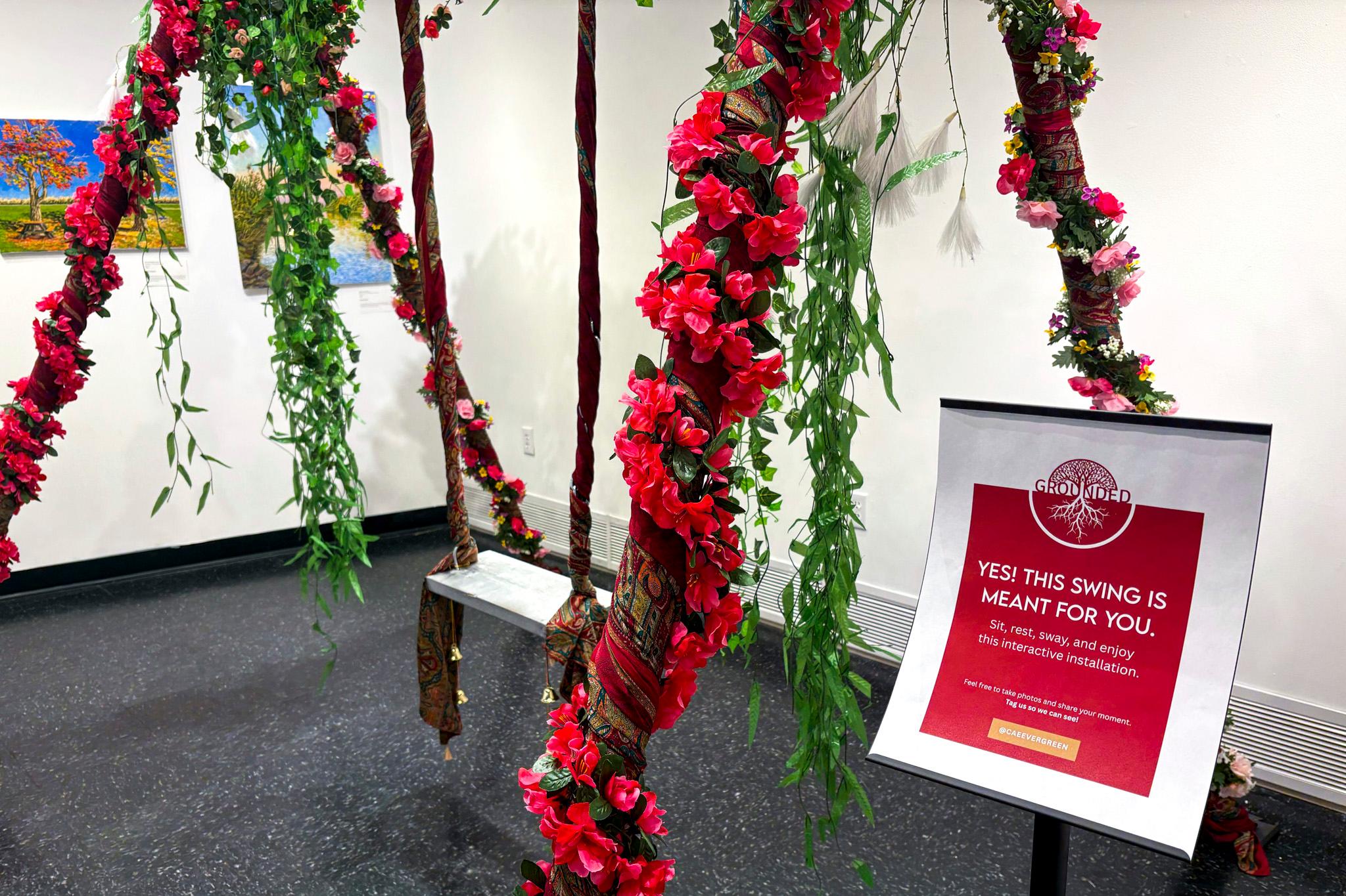 A swing is suspended from a ceiling in a white room, draped with pink flowers and green vines. A sign in the foreground reads "YES! THIS SWING IS MEANT FOR YOU."