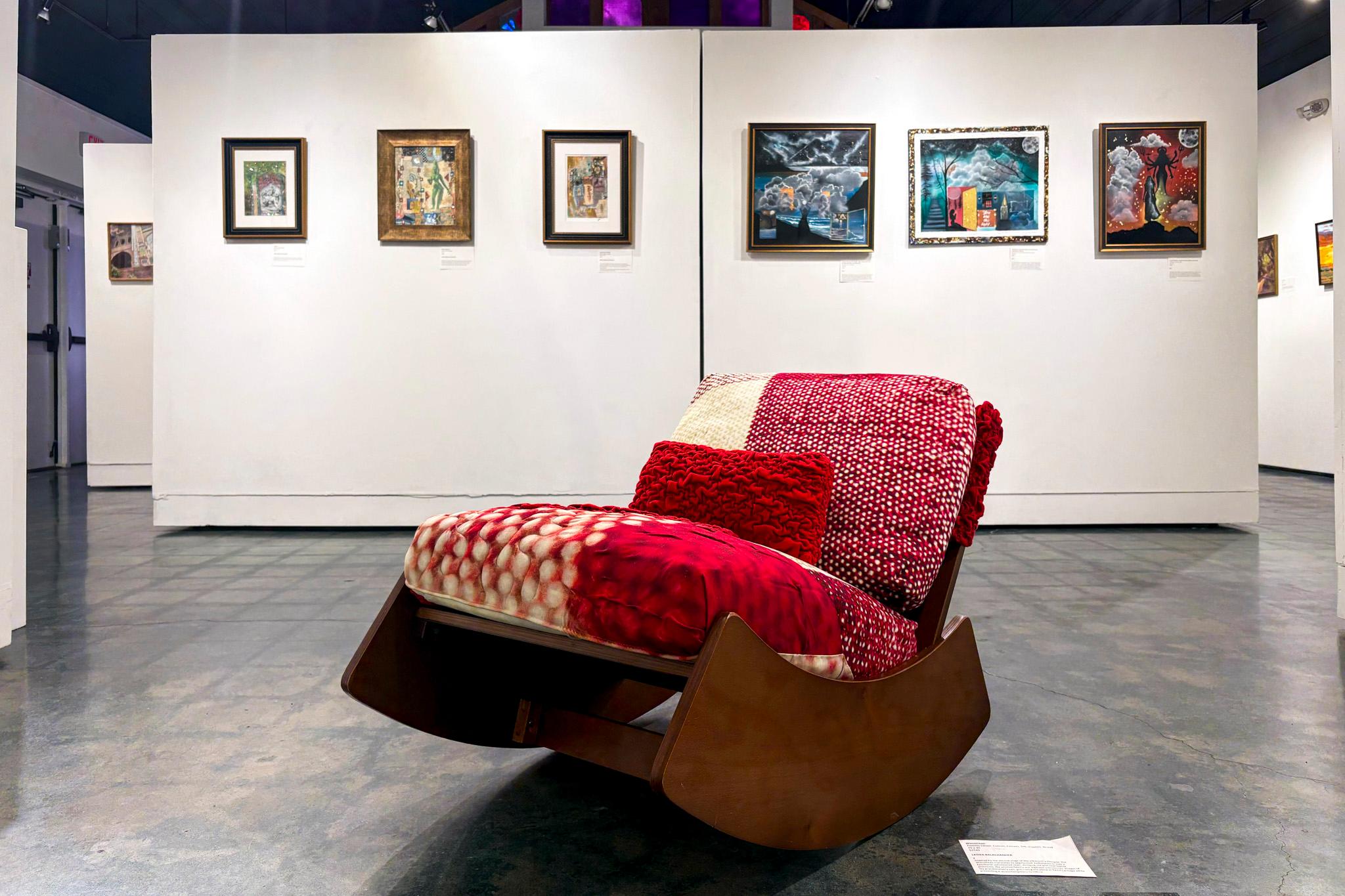 A mod rocking chair, covered with red and white upholstery, sits on a slate floor with paintings mounted to the walls behind it.