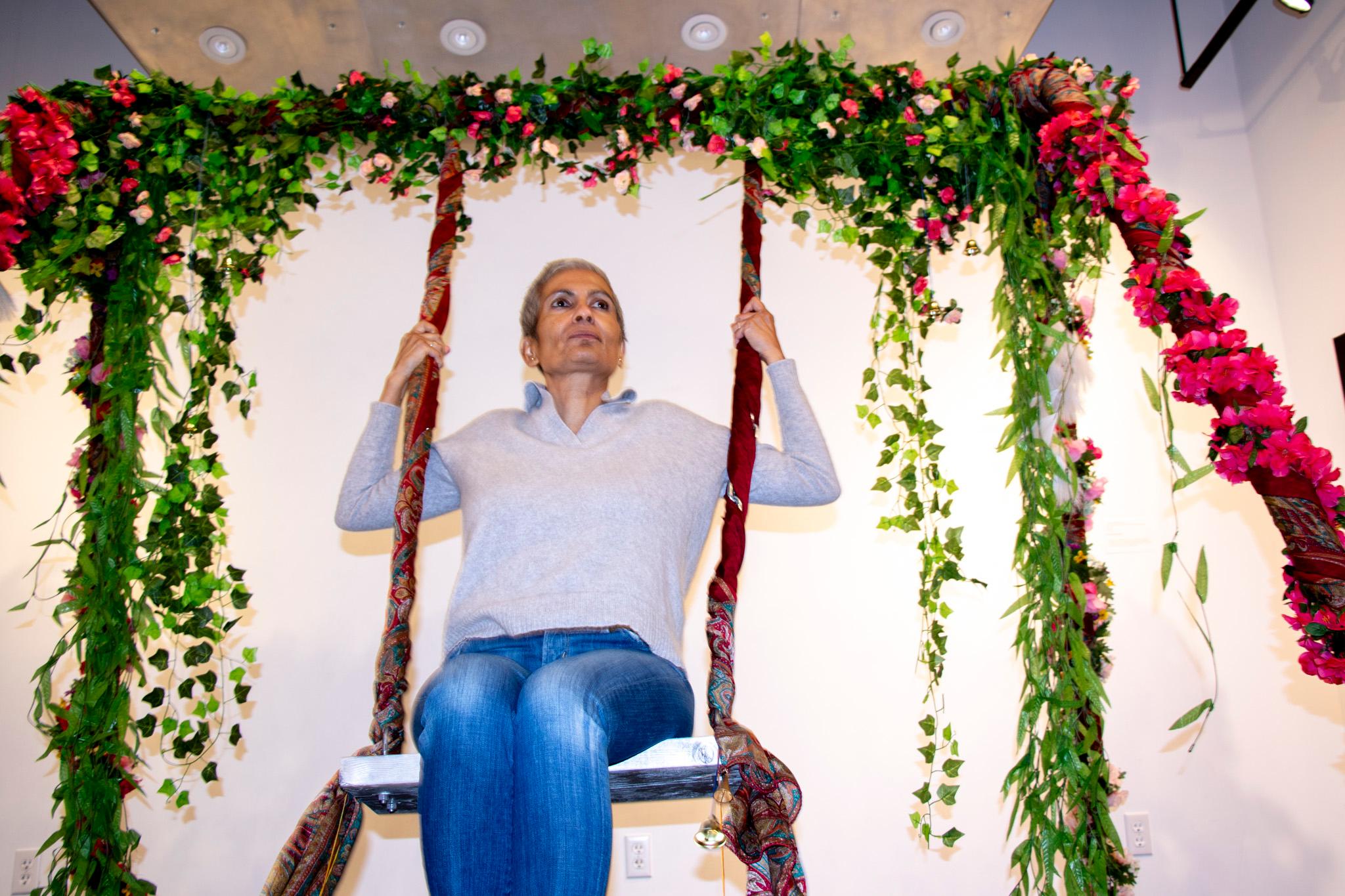 A woman with short hair and a collared shirt rides a swing built in a white room. Pink flowers and green leaves hang off its frame.