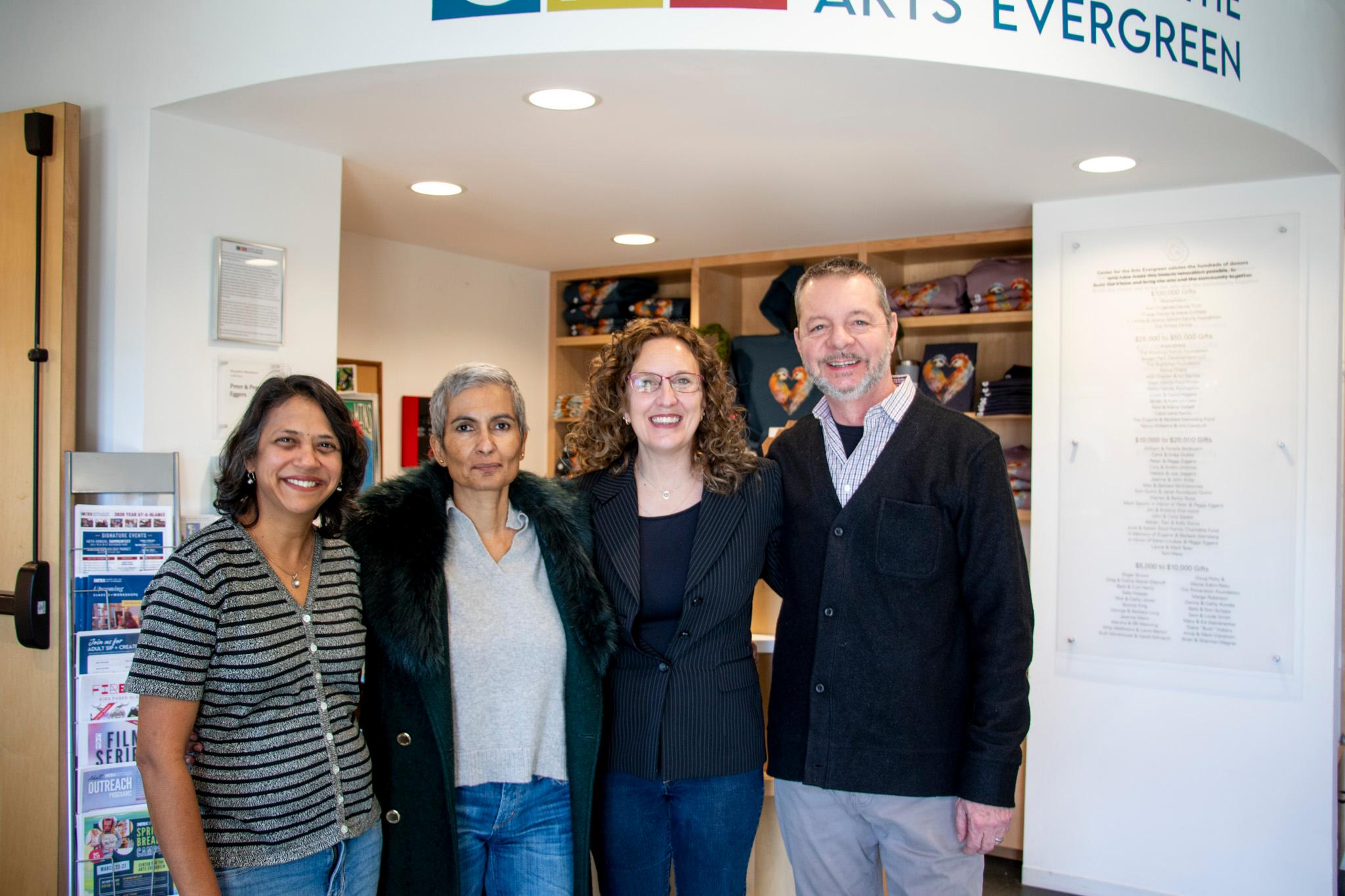 Four people stand together, arm in arm, smiling at the camera.