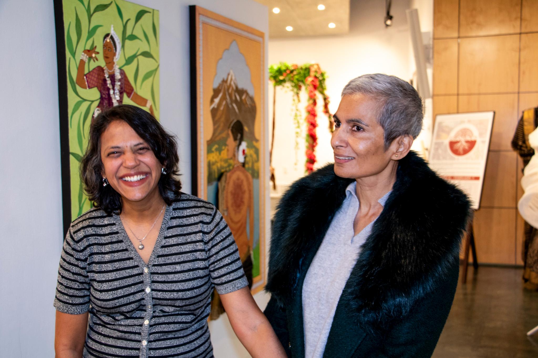 Two women, one wearing a fuzzy black coat, stand in a warmly lit room with art hanging on the walls.
