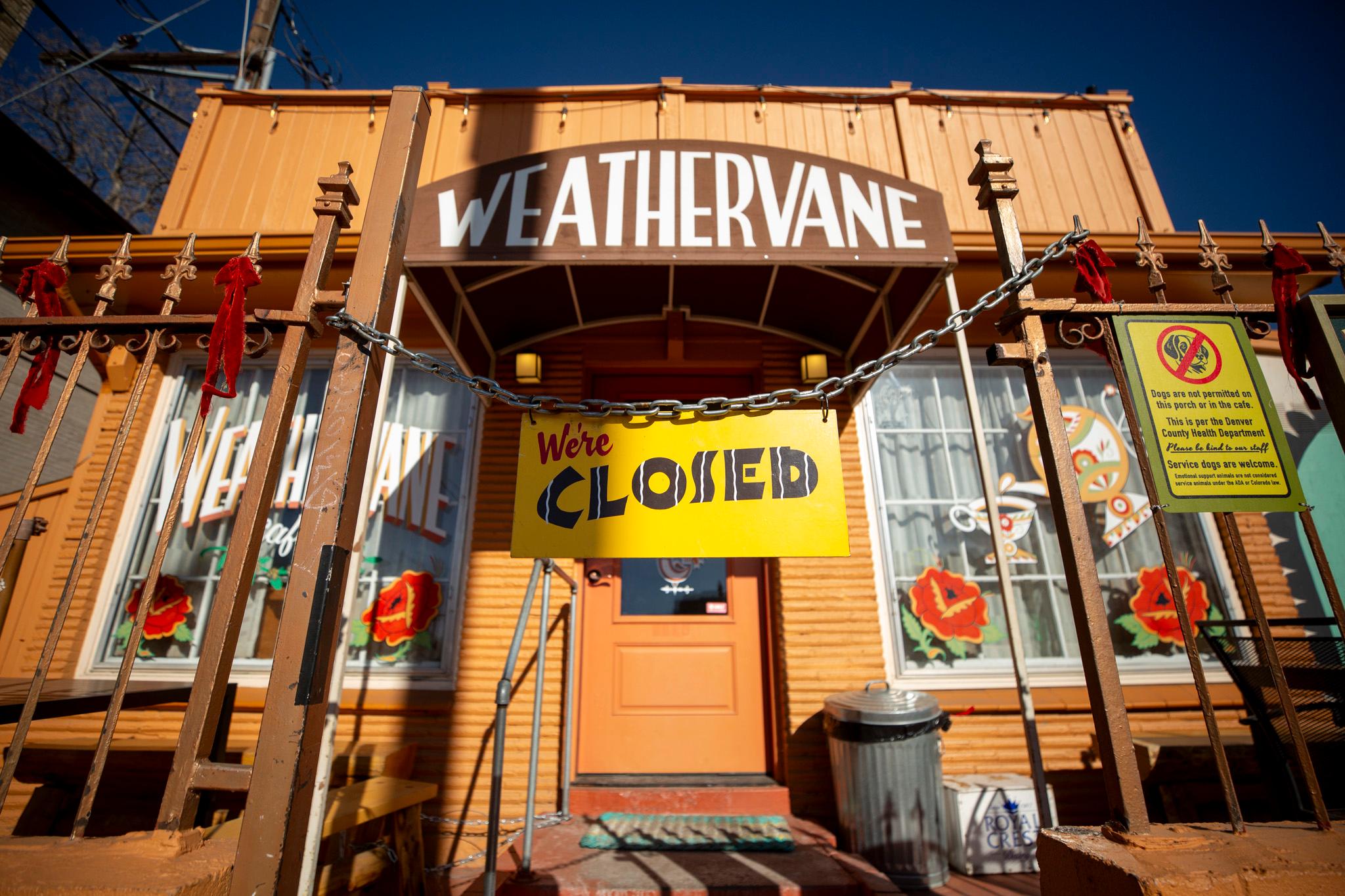 The front of a light brown building is seen through an opening in a fence that is chained off with a yellow sign reading "We're closed."