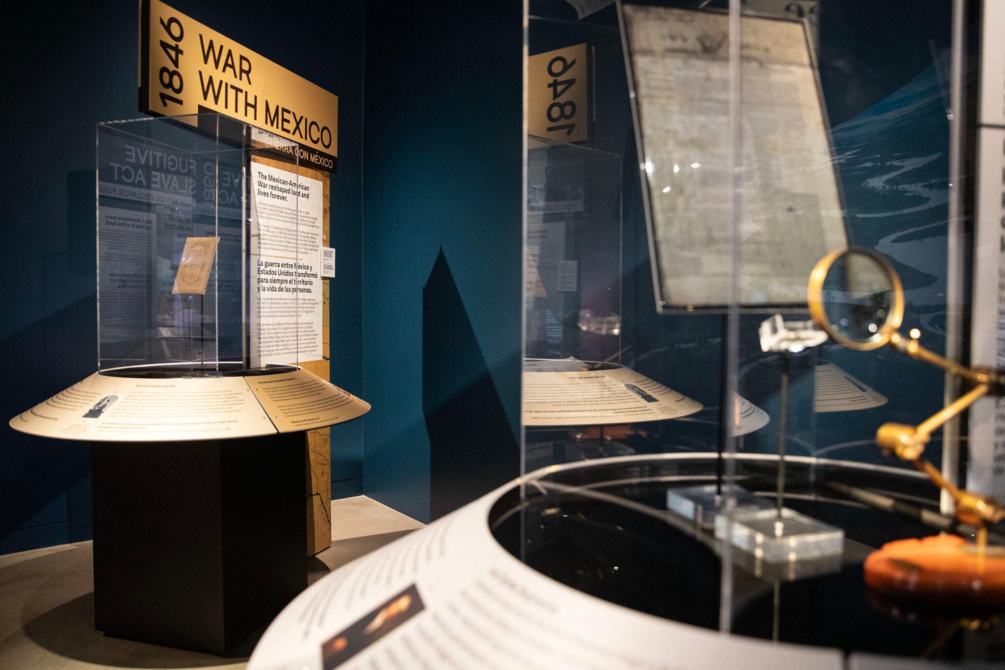 Museum display cases in a blue room. They're filled with printed material and at least one magnifying glass.