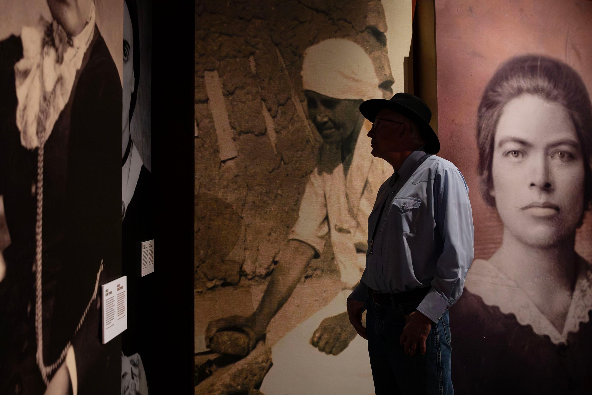 A man in a brimmed hat is silhouetted in a room covered with black-and-white photographs of women in old-timey clothing.
