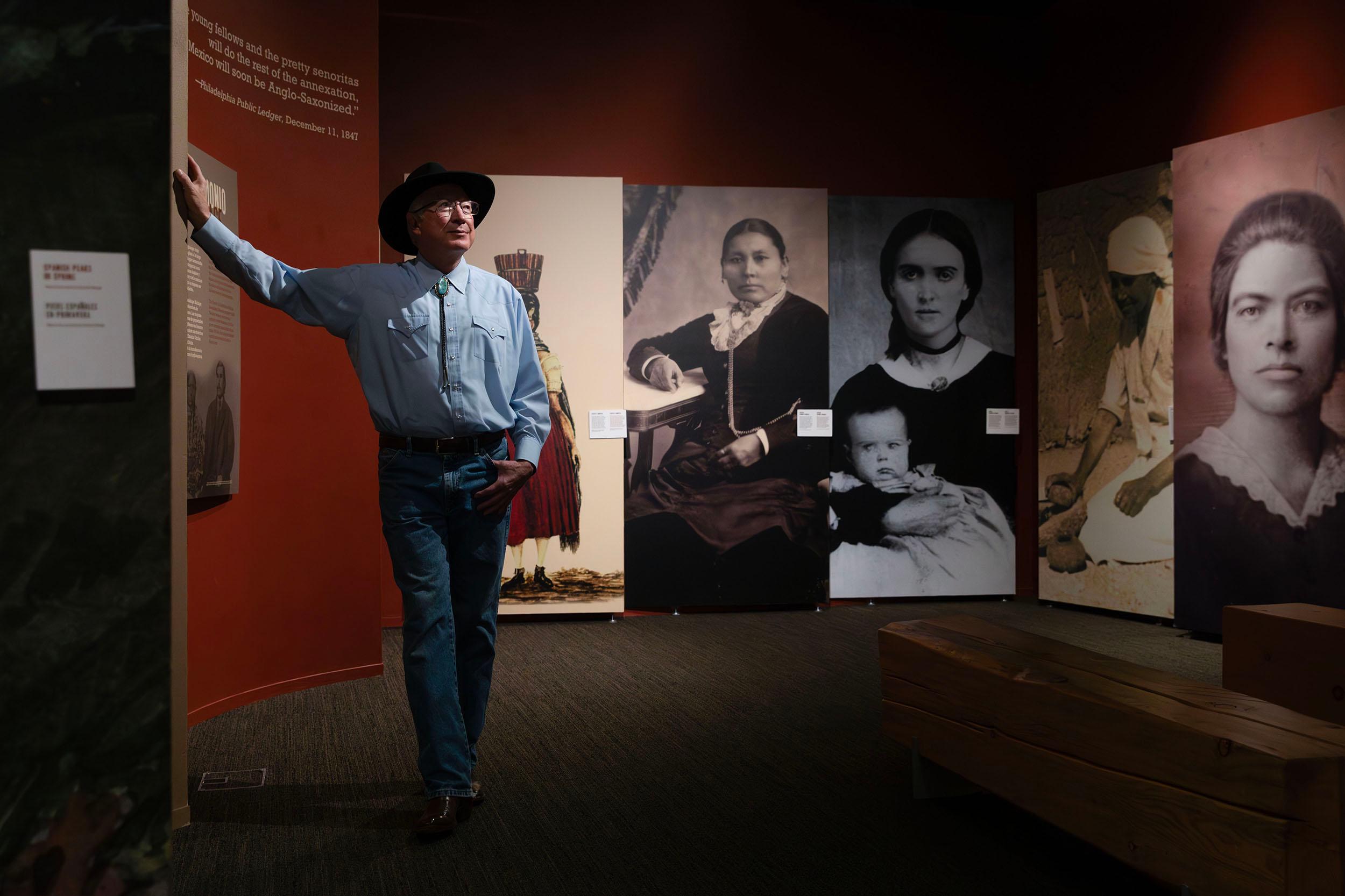 A man in a wide-brimmed hat and a turquoise bolo tie leans on a wall in a maroon room covered with large black and white photographs of women wearing old-timey clothing.