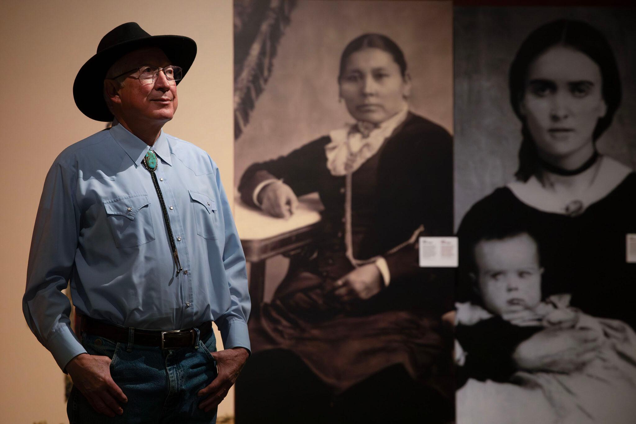 A man in a wide-brimmed hat and a turquoise bolo tie stands before large black and white photographs of women wearing old-timey clothing.