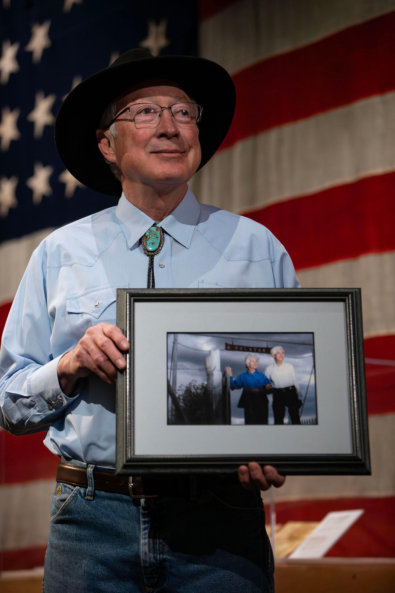A man in a brimmed hat stands before a giant American flag, holding a photograph of two people with white hair.