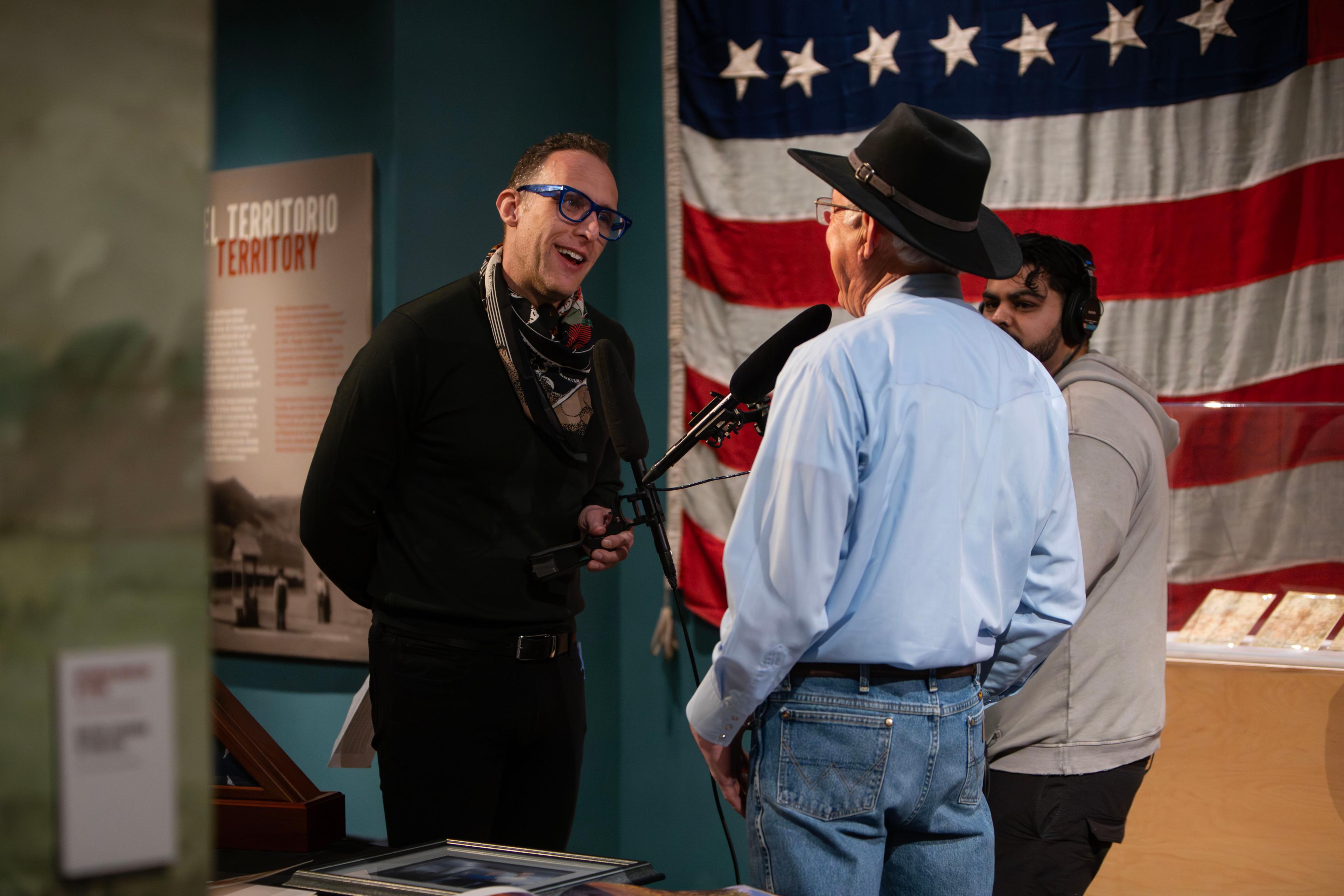 A bespectacled man in a dark shirt smiles as he points a microphone towards another man in a wide-brimmed hat. An American flag hangs on the wall behind them.