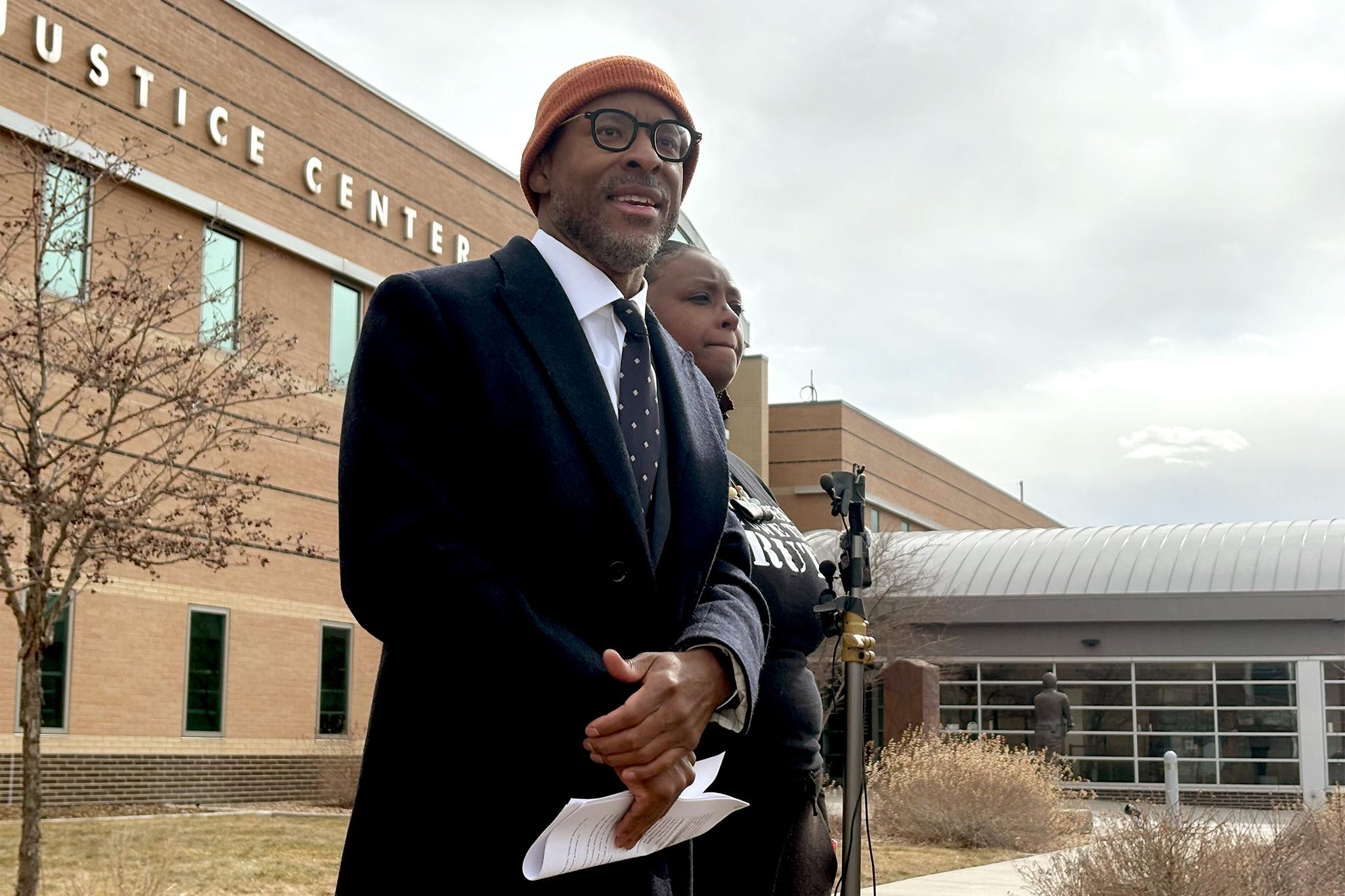 A man in a black suit, glasses and a beanie speaks next to a stand with microphones clipped to it. A woman stands just behind him, pursing her lips. A tan brick building rises behind them.