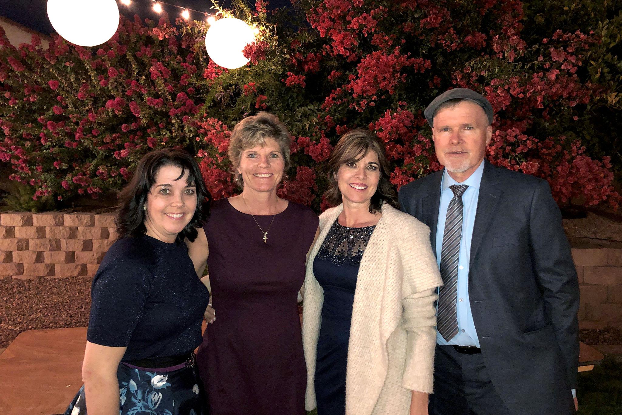 Three women and a man wear nice clothing and stand in front of a pink flowering bush.