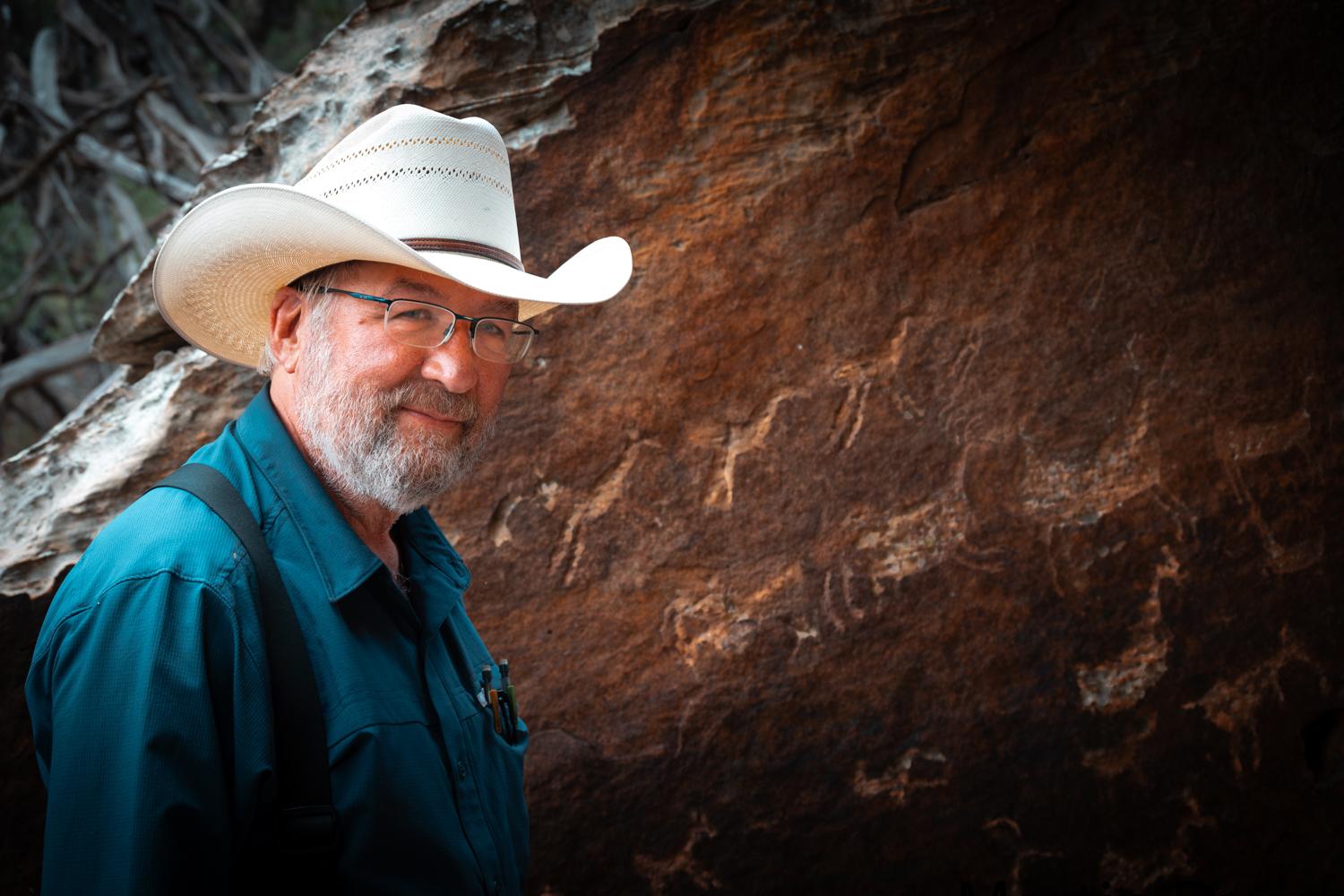 A man wearing a cowboy hat, suspenders and glasses is standing in front of a rock with carvings on it.