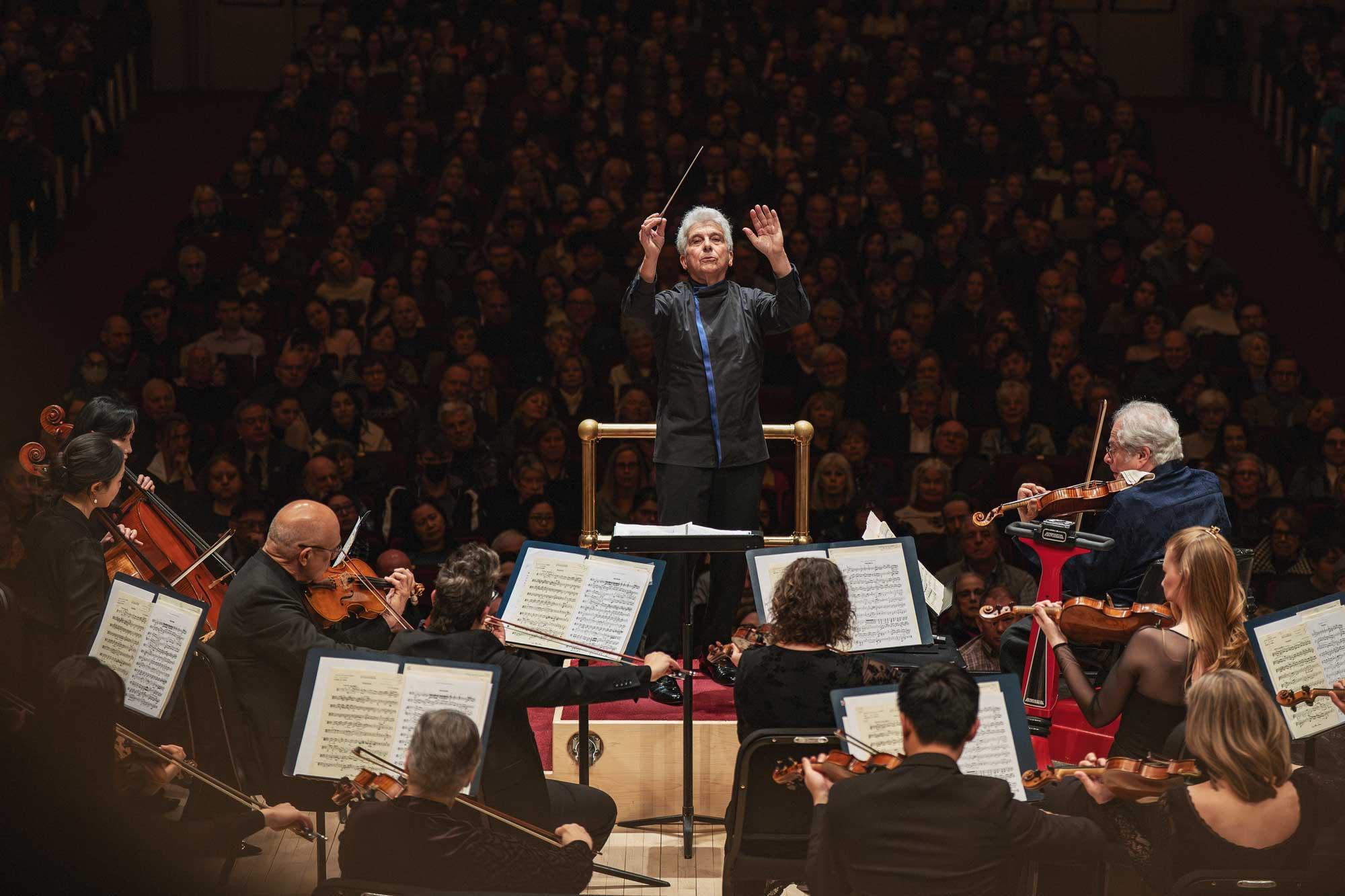 An older man faces the camera, conducting an orchestra. A large crowd sits in dark theater seats behind him.