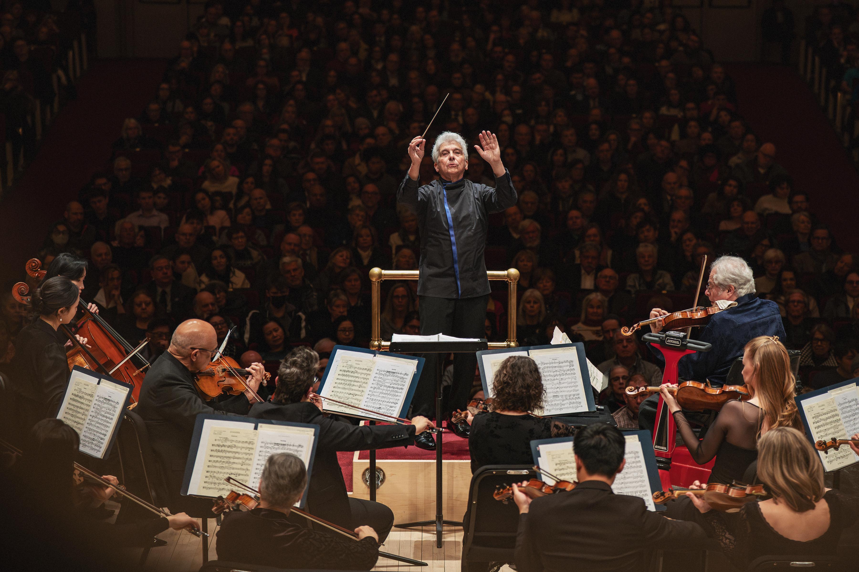 An older man faces the camera, conducting an orchestra. A large crowd sits in dark theater seats behind him.