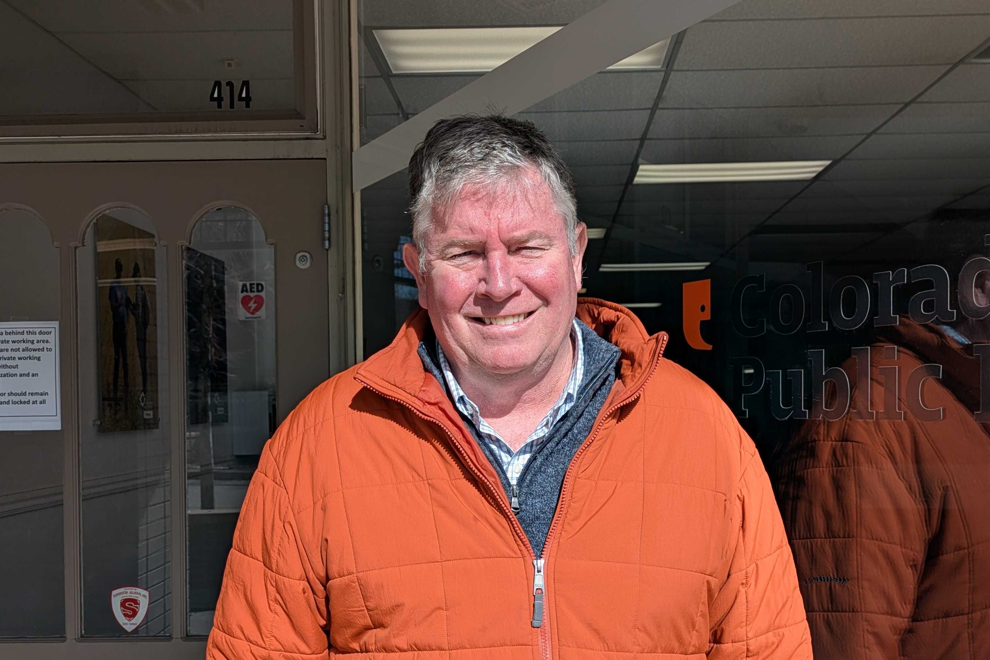 Image shows an older man with white hair wearing an orange winter coat