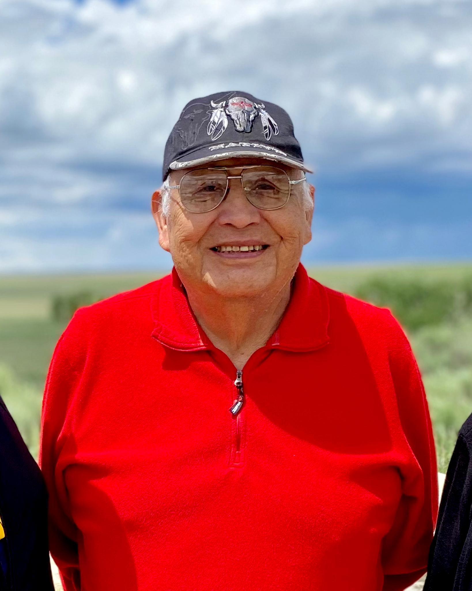 A older Native American man wears a red shirt, rimmed glasses, and a black hat.