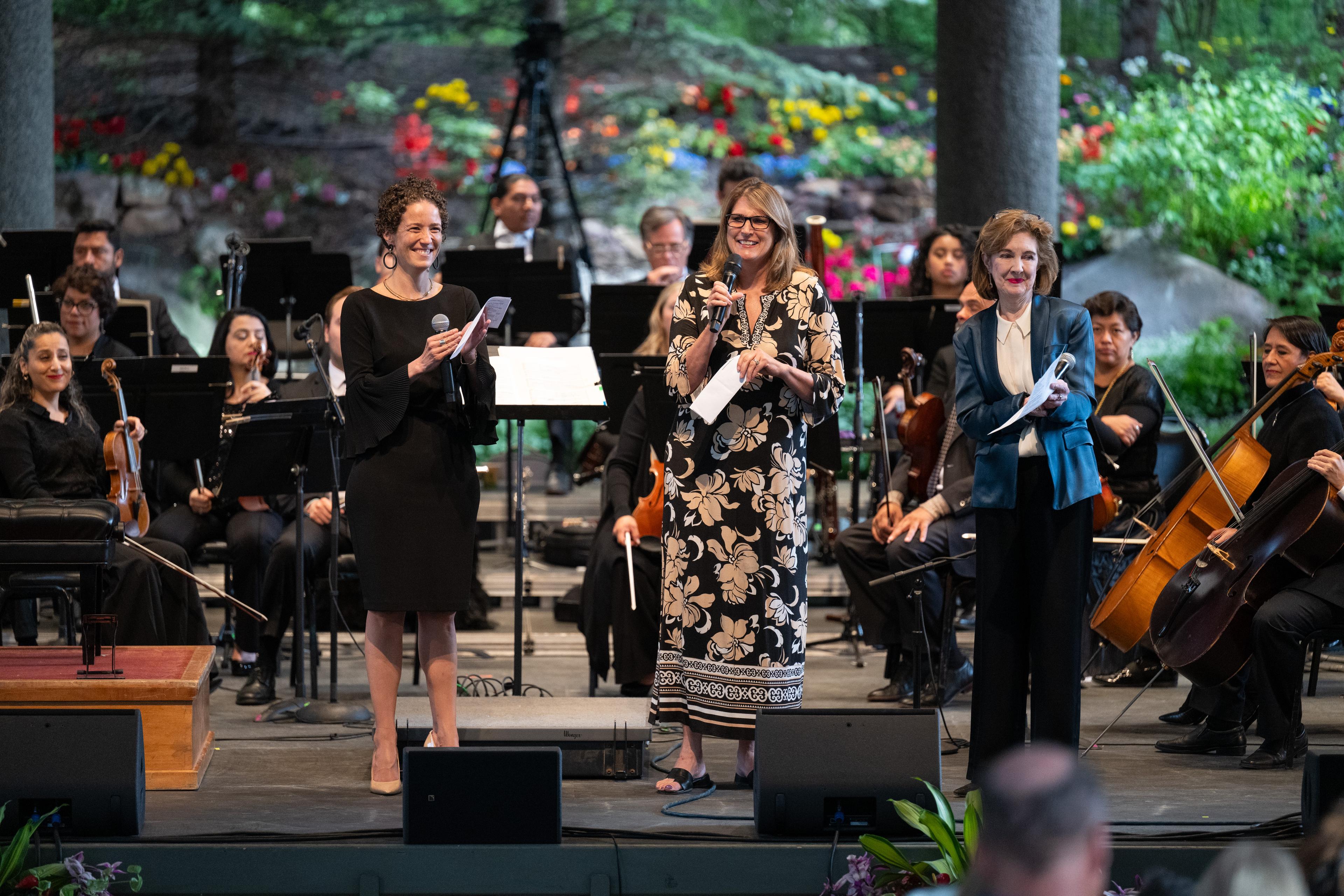 Three women standing in front of an orchestra talking to the audience.