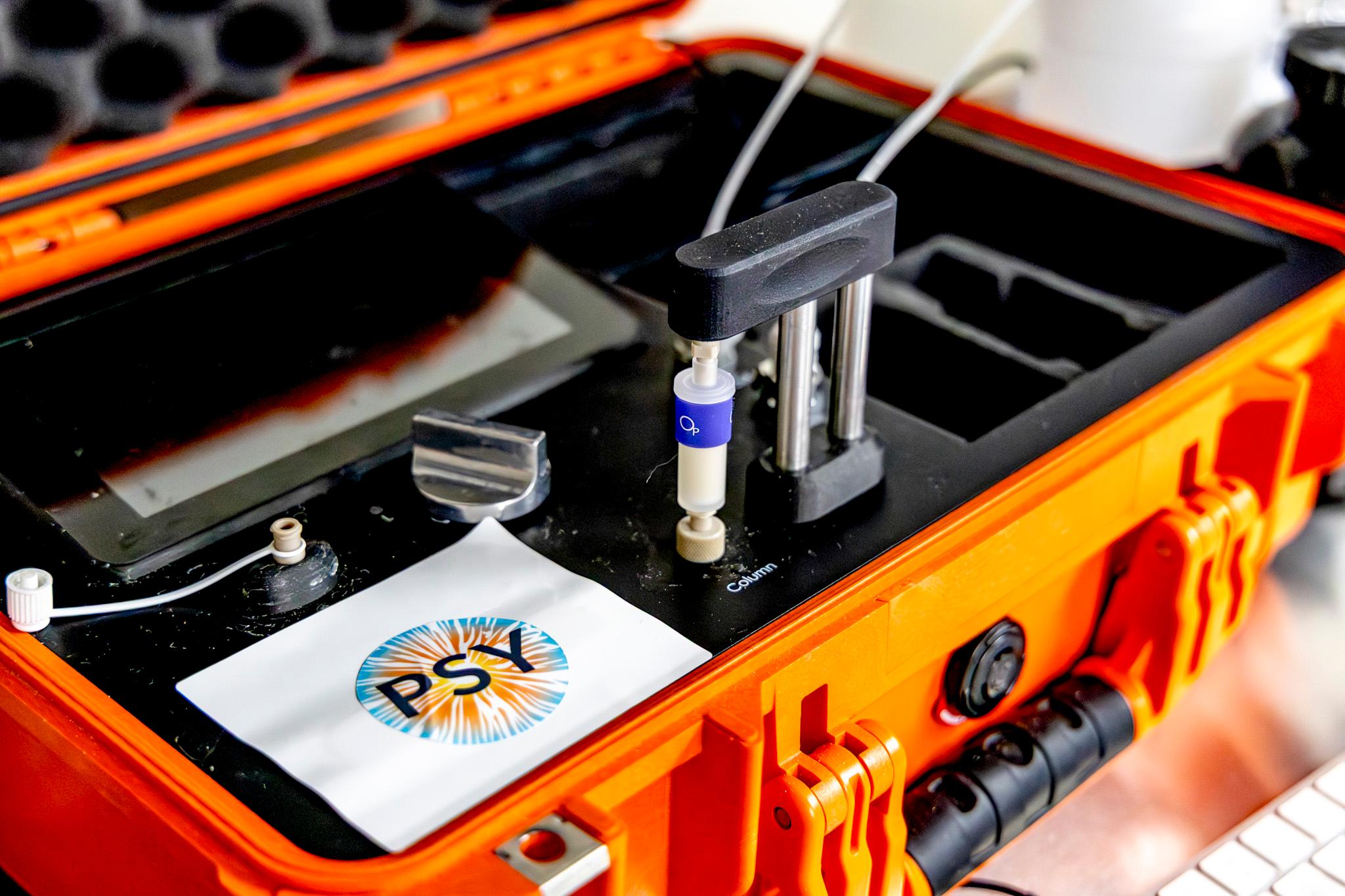 An orange case contains black and metal equipment. A sticker on top reads "PSY."