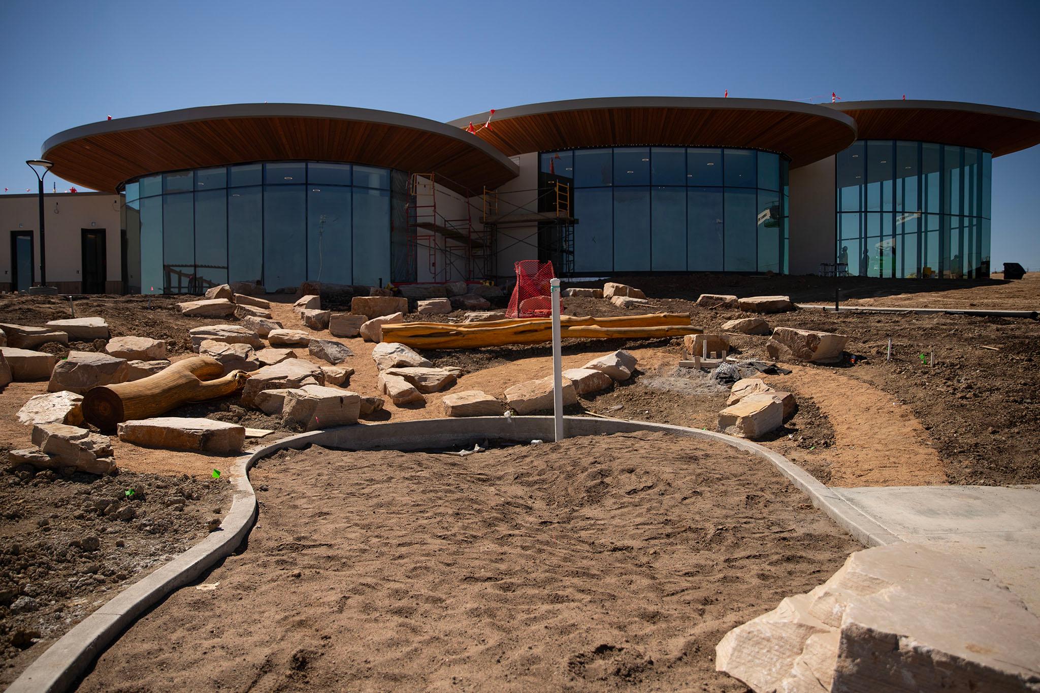 A building topped with a curving, wooden roof and tall glass windows sits on a dirt lot.