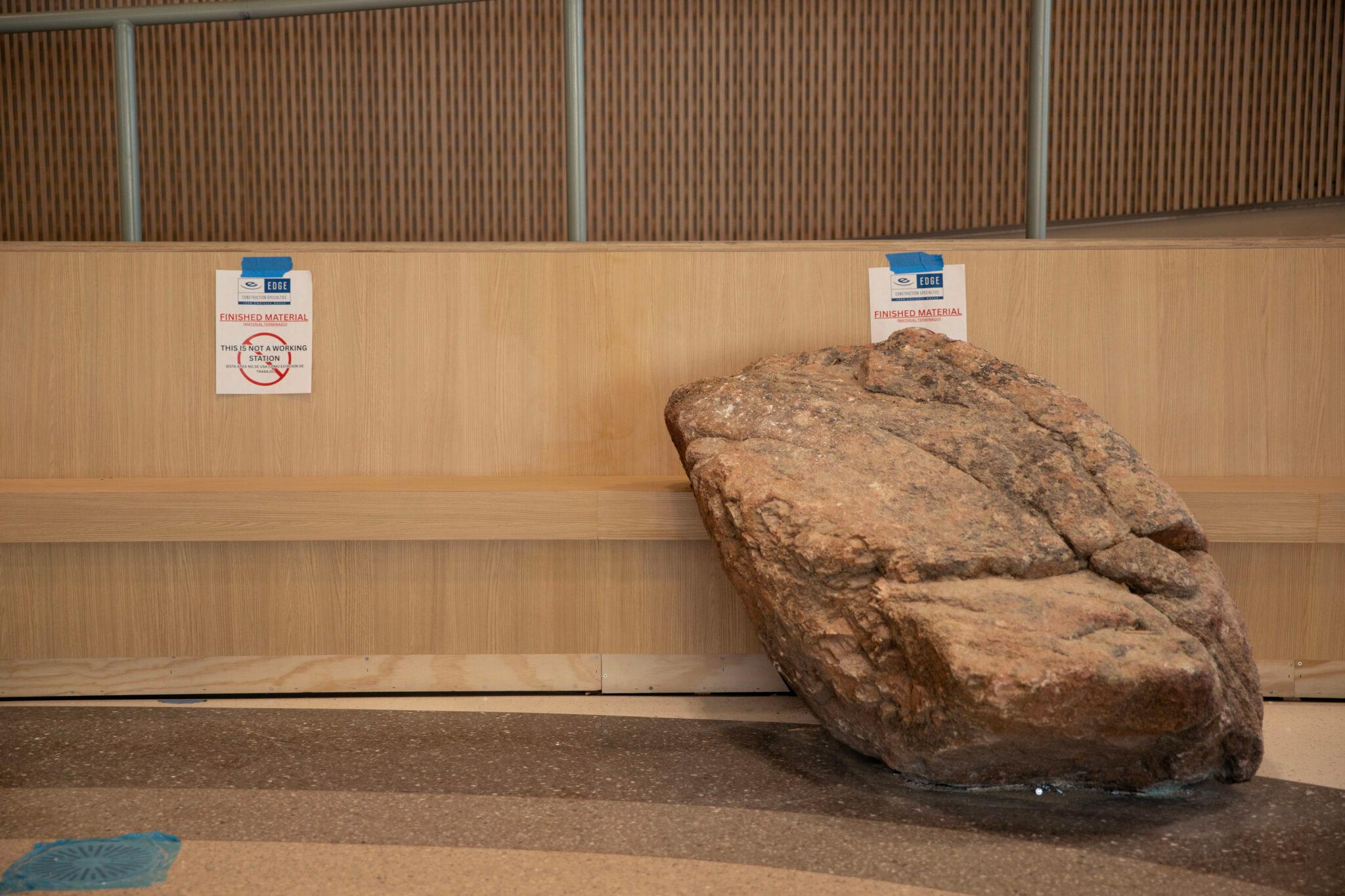 A large boulder the size of a small boulder sits by wooden benches in a beige room.