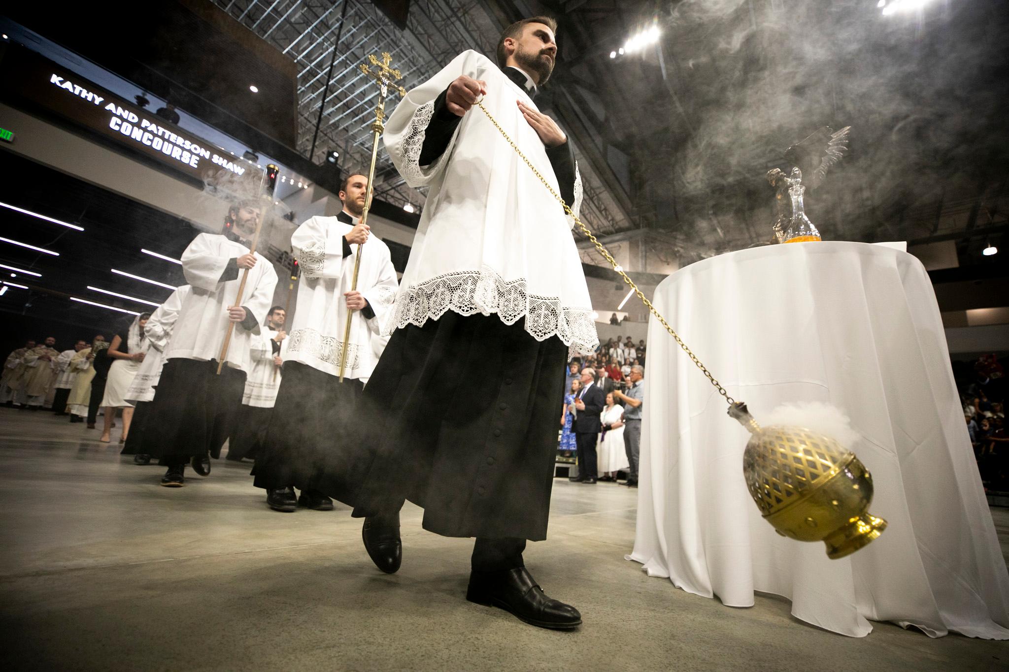 A man in a white frock swings a golden chain — at the end of it is a golden, metal acorn emanating smoke.