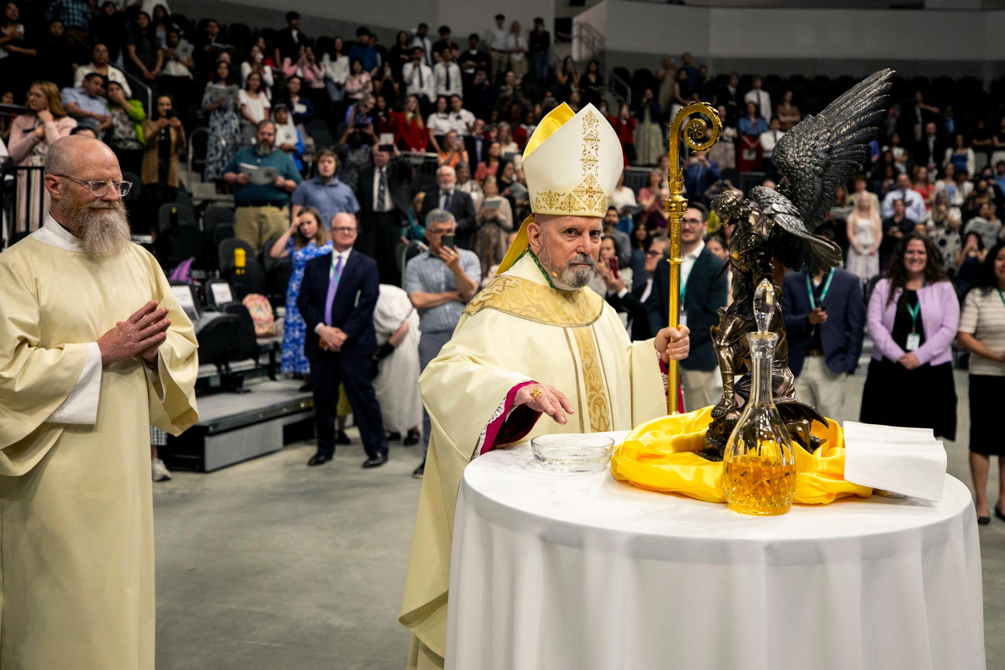 A man in gold robes and a pointy fish hat — and holding a staff with a curling top — looks right at the camera as he dips his fingers into a bowl of water. Behind him is an arena full of people.