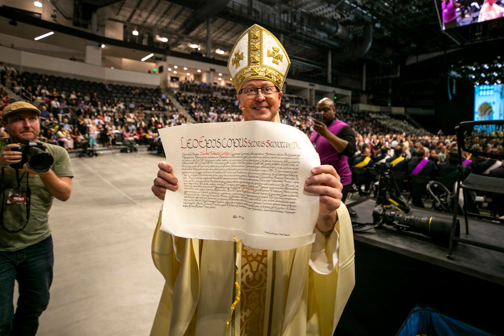 A man in an ornate, golden fish hat smiles as he holds up a sheet of paper to the camera covered in intricate script.