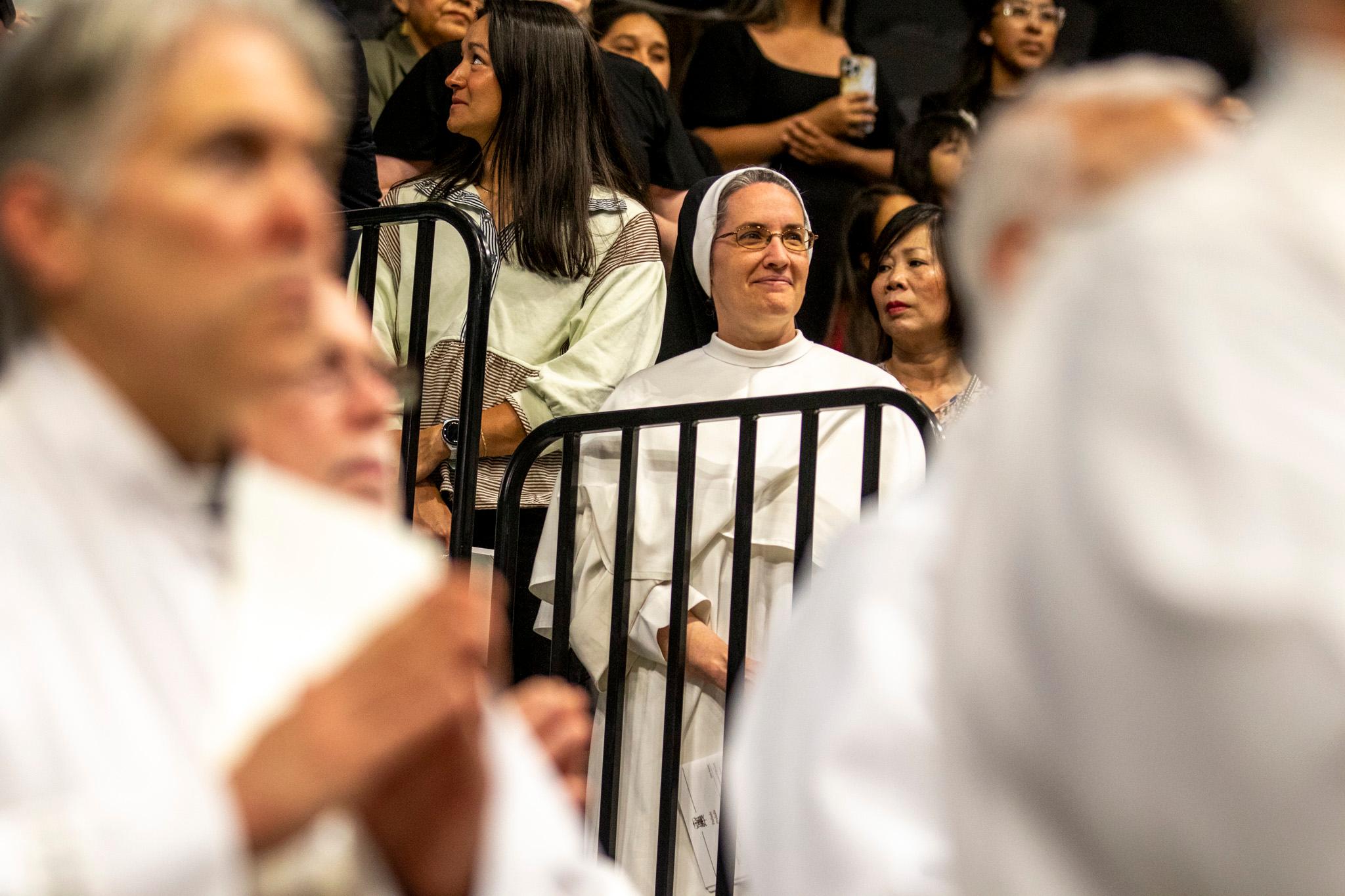 A woman in a white robe and black-and-white nun hat smiles as men in white robes walk past before her.