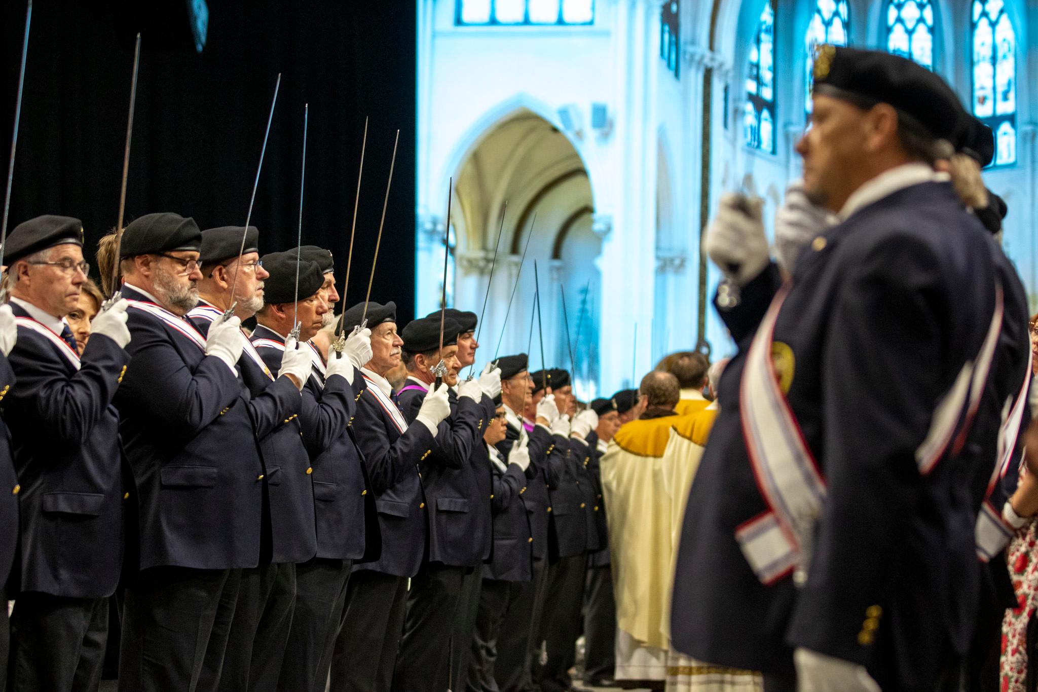 Men in blue suits, wearing black berets, hold thin swords in front of their faces in two rows.