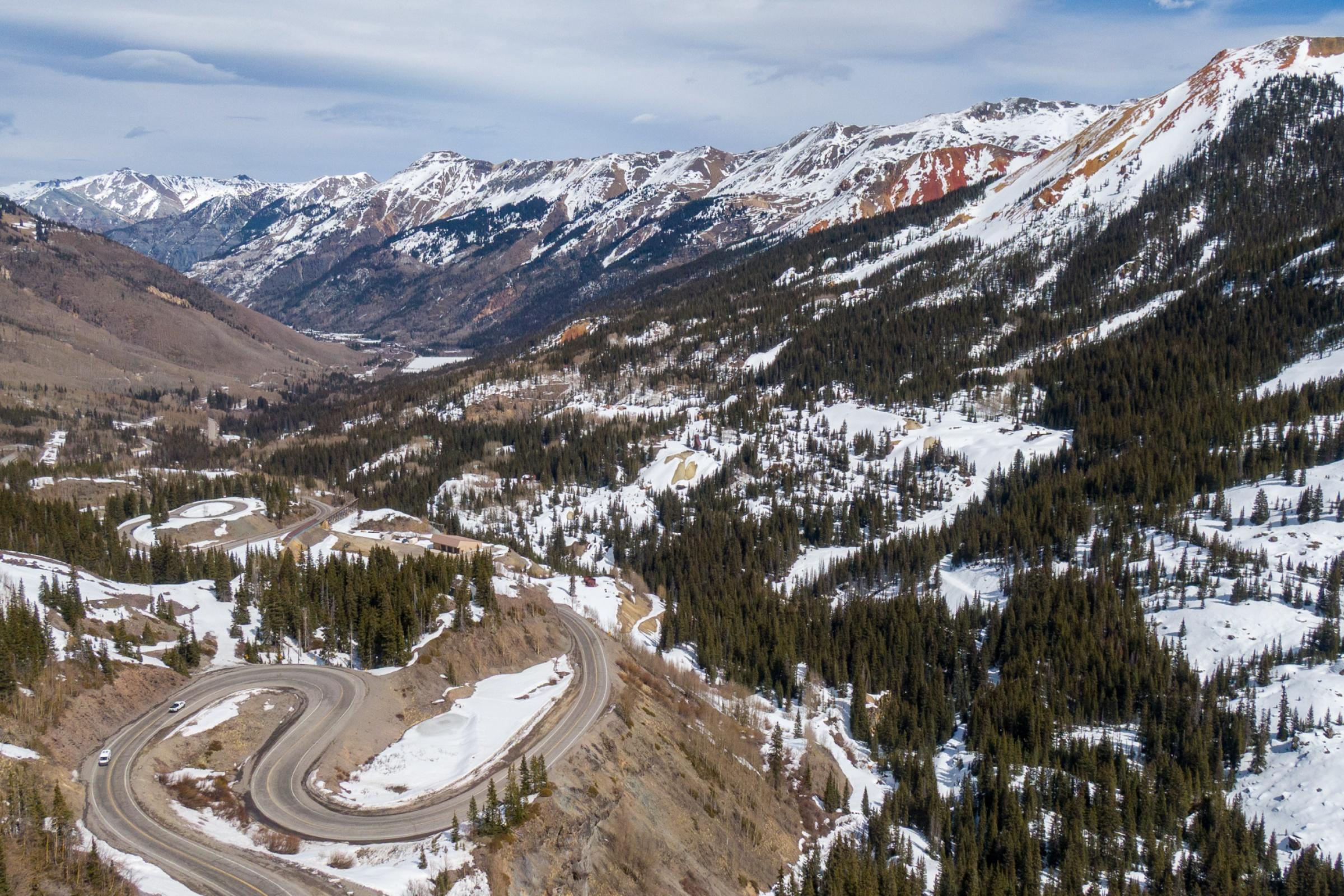 an aerial shot of a road with mountains with some snow in the background