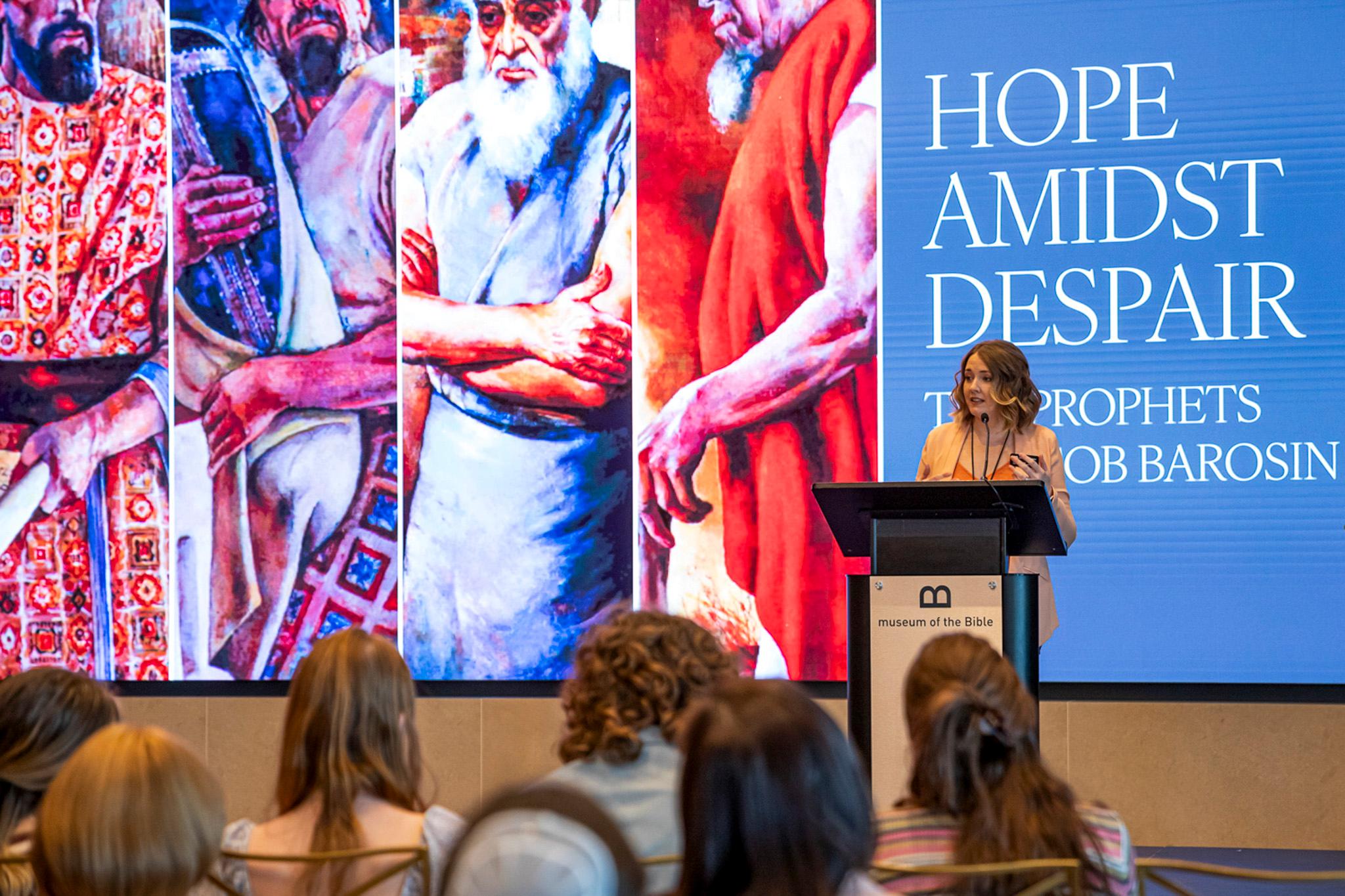 a woman speaks behind a lecturn. in the background are colorful images and text