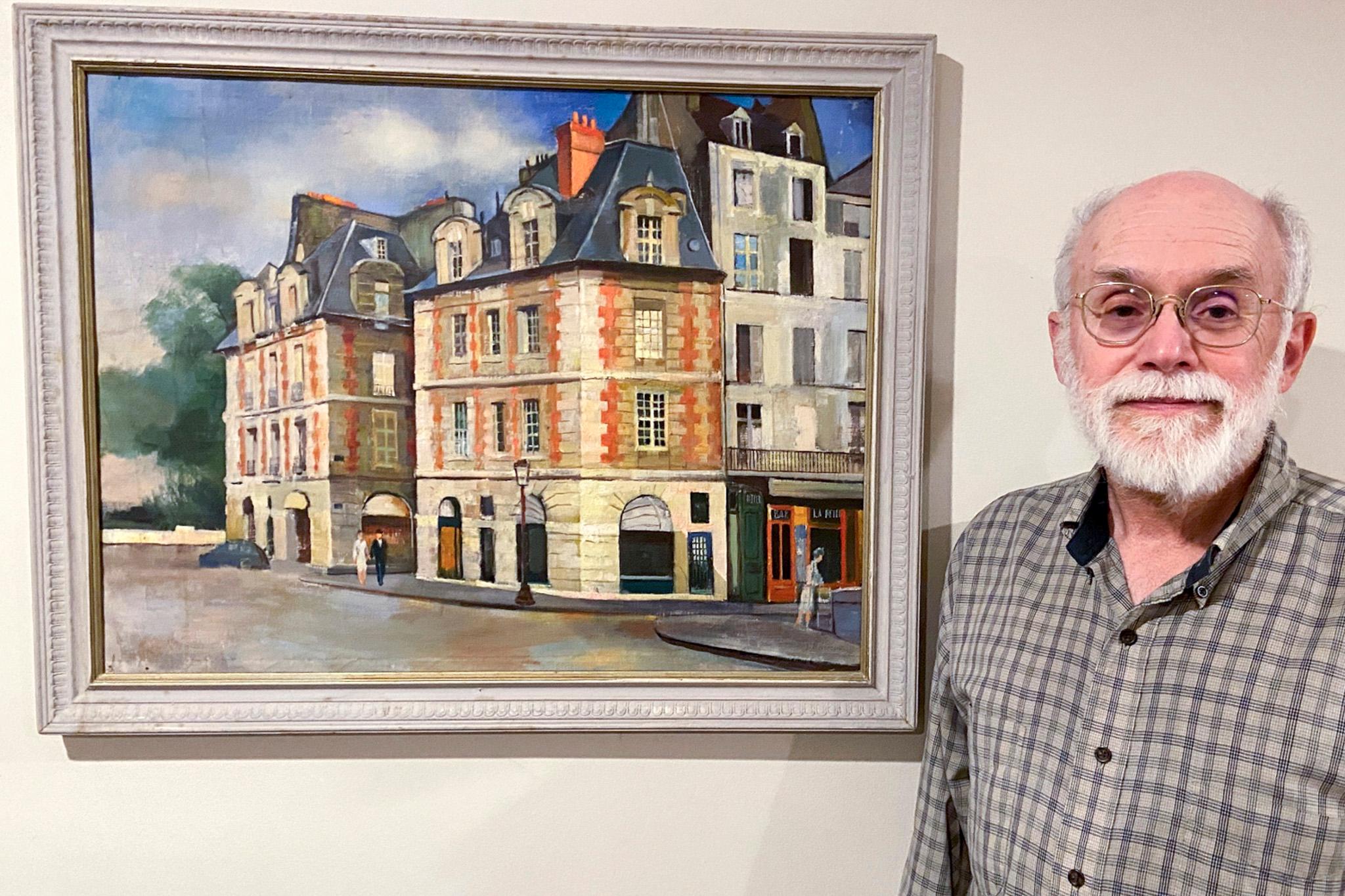 a man with glasses stands in front of a framed painting of buildings