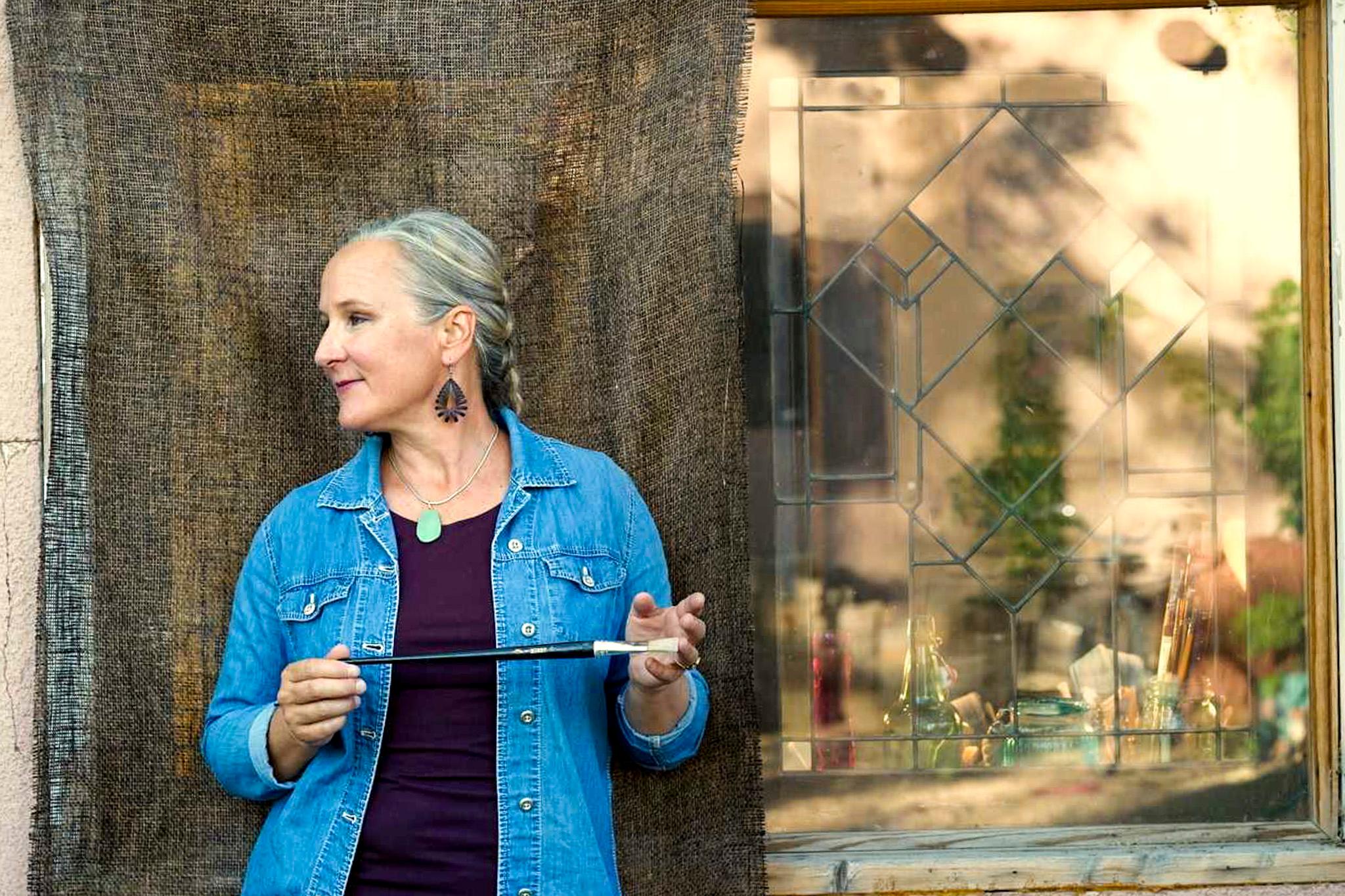 a woman holds a paint brush. She stands next to a window where art supplies can be seen inside