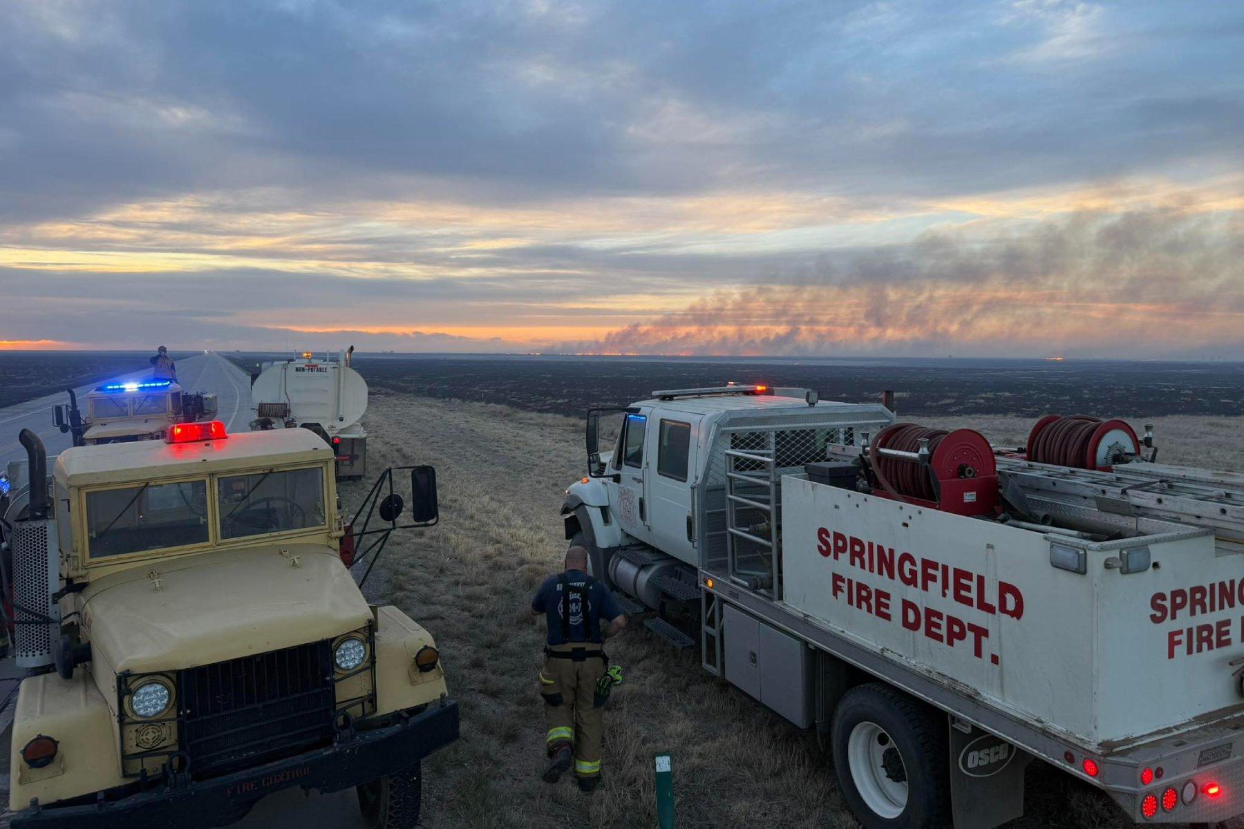 Two trucks, one with "Springfield Fire Dept." on the side are in the foreground of a gravel road as smoke billows from a fire far on the horizon. It's sunset.