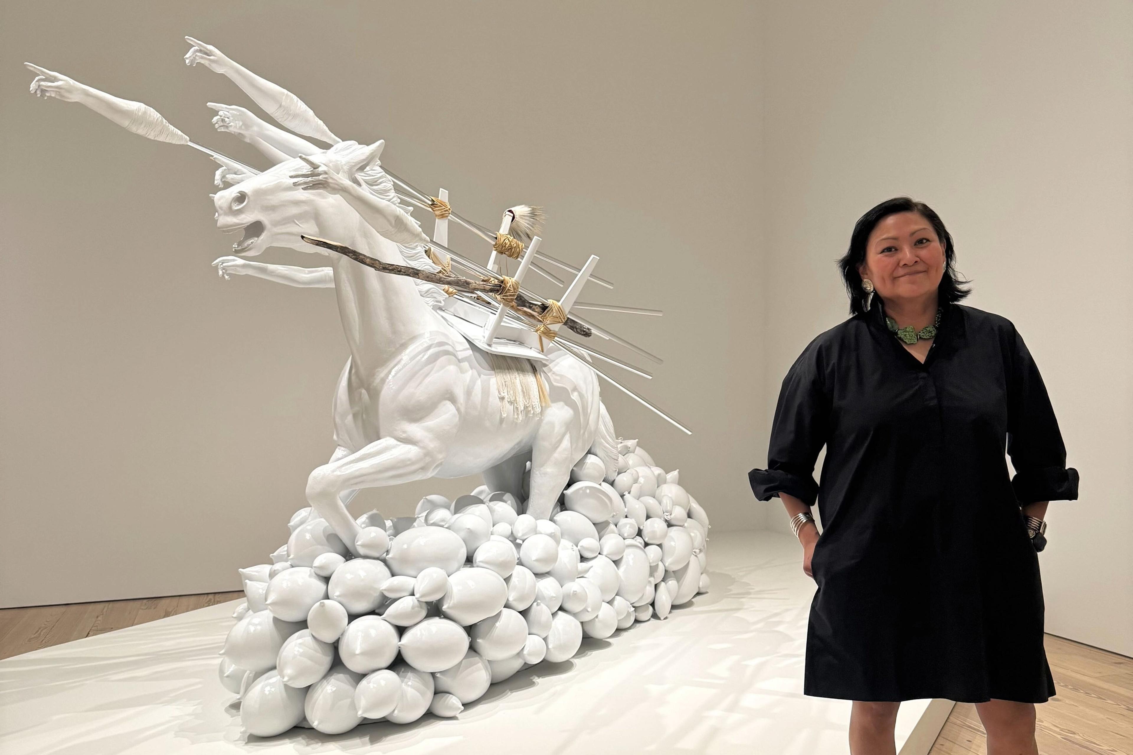 Woman in a black dress stands on the right next to a larger than life white sculpture of a horse embellished with castings of arms and other objects.