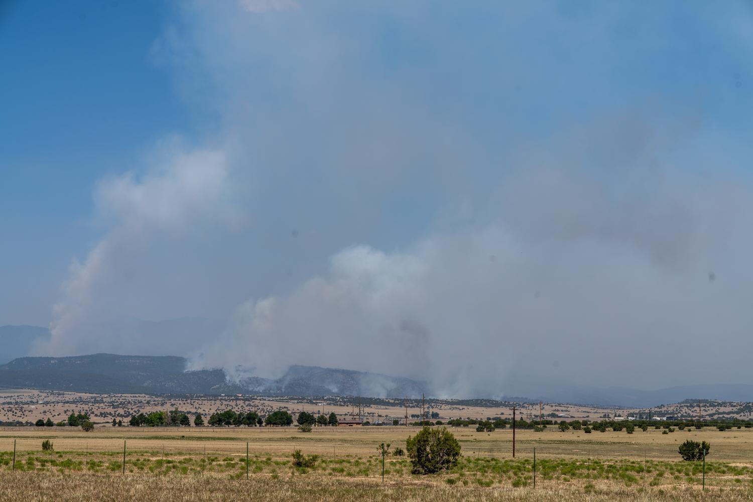 clouds of smoke rise in the distance from forested hills